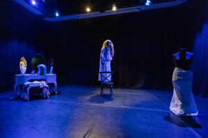 1. Blue light. A view of the whole stage with, left to right, a dressing table and triptych mirrors, a woman wearing a simple white dress and white headband over her long loose hair standing on a chair looking warily at the audience and a black tailor’s dummy with no arms or head, its lower-half wrapped in white cloth.