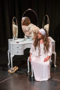 1. The same woman in the same costume sits on a stool in front of the dresser, looking at the audience to her right. She is reflected in the mirrors.